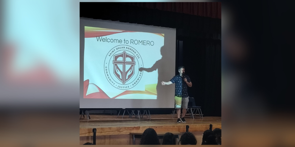 A person on a school stage in front of a projector that says "Welcome to ROMERO"