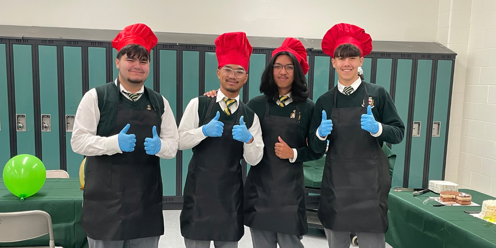 Four chefs wearing red tall hats and black aprons stand in a locker-room setting, smiling and giving thumbs-up with blue gloves; green lockers, balloons, and a table with food are in the background.