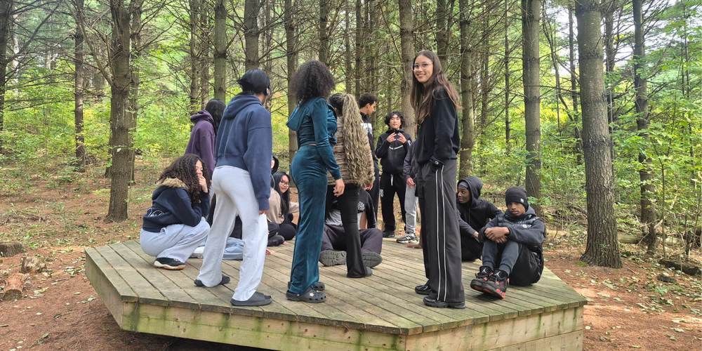 Group of people standing and sitting on a raised wooden platform in a forest, with backpacks and outdoor clothing, surrounded by tall trees and a dirt clearing.