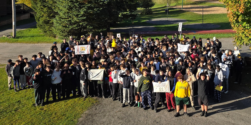Group photo of Chaminade students and staff at Camp Manitouon a sunny autumn day, holding signs before colorful fall trees