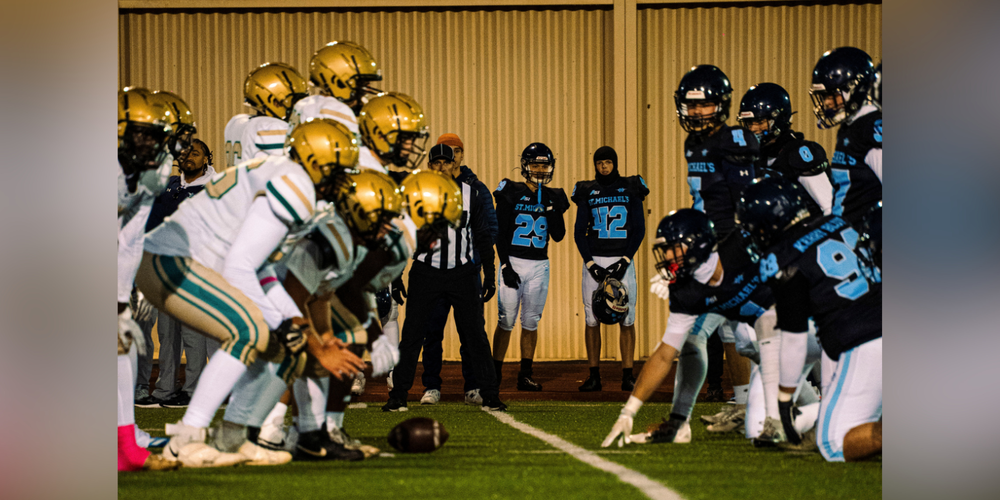 Football teams lined up at field level, with players in gold and white helmets facing players in dark blue uniforms and helmets during a game near a brick wall.