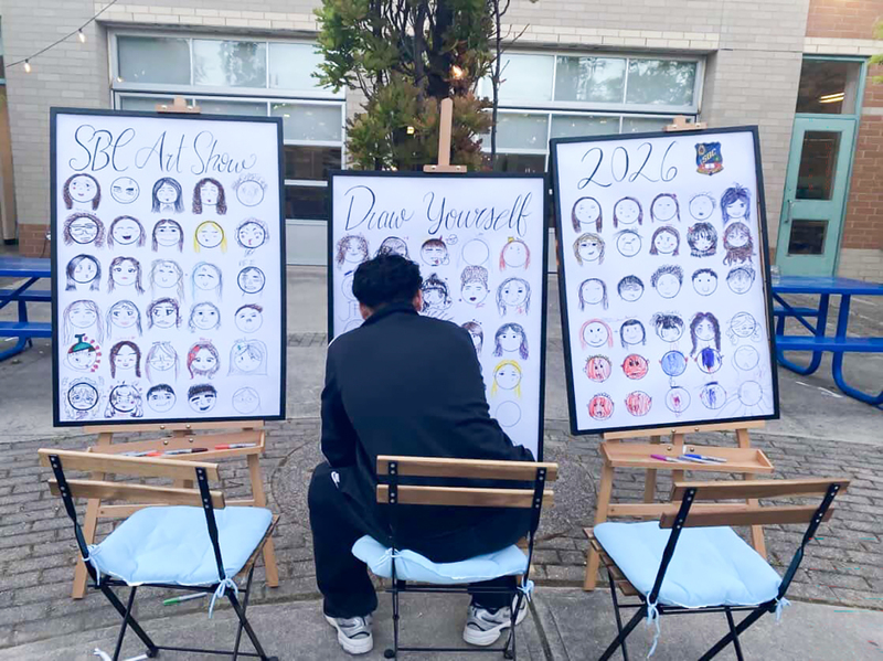 Photo of a student drawing themselves at the Draw Yourself section where there are three boards filled with students' protraits of themselves