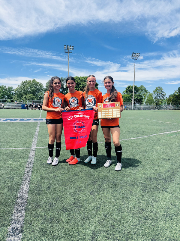 Photo of four members of the St. Cecilia soccer team posing on the field with their championship banner and plaque