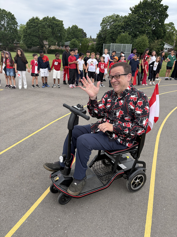 Photo of St. John Viannery students in the school grounds holding hands and wearing Canada shirts, along with a staff member in the foreground on a scooter with a Canada flag on the scooter and wearing a Canada shirt