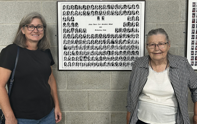 Photo of Deb Smith and Olga Hilmer standing in front of the graduating class plaque that has Father Hilmer's graduating photo.