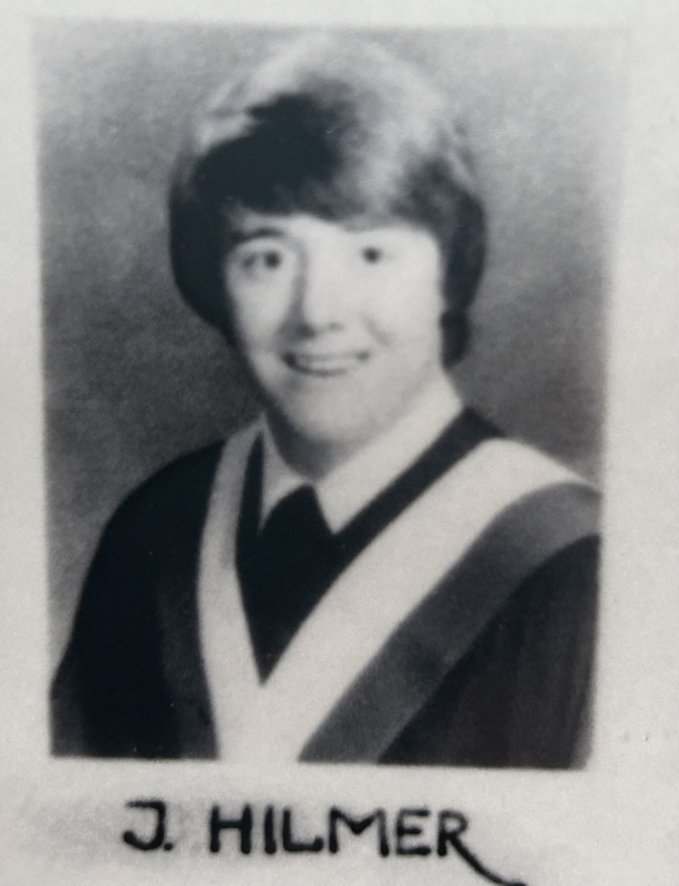 Father Hilmer's high school graduating photo in black and white with the text J. Hilmer below, showing a young man in graduating robes smiling at the camera