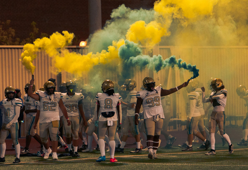 Photo of Chaminade football student athletes in full gear on the court, setting out colorful plumes of smoke in the Chaminade colours of yellow and green
