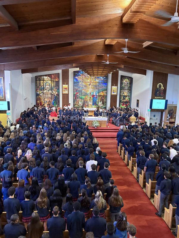 Redmond students and staff attend the Mass in the church, facing the spakers with huge sweeping religious stained glass mural artwork beyond the speakers