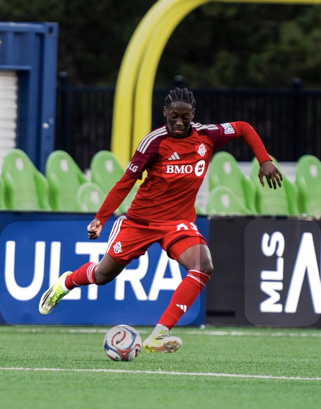 Edwin, a Toronto FC player in a red kit runs toward a soccer ball on the field, poised to kick, with stadium seats and MLS signage behind him.