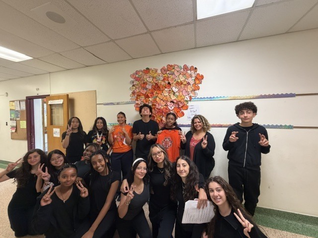 A group of students and teachers pose with peace signs in a school hallway beneath a colorful display of a heart made of orange hearts with messages written on them