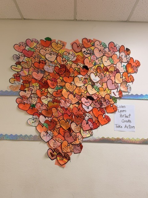 A heart-shaped collage of colorful paper hearts, covered in handwritten words and drawings, decorates a classroom wall beside a “Listen, Reflect, Create, Take Action” sign