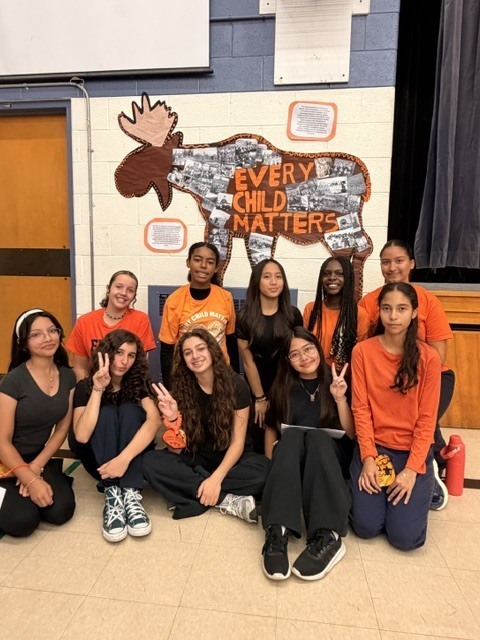 OLW students pose beneath a collage art of a moose reading “Every Child Matters,” the students wearing orange and black in a warm, respectful school display.