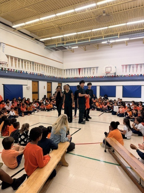 Students sit on gym benches around several students performing in the center of a school assembly, creating an attentive, lively atmosphere.