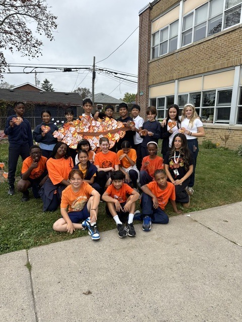 A cheerful group of students in orange shirts pose outside a school, holding a collage art made of orange hearts with messages written on them, in the shape of a feather, beside the school brick building.