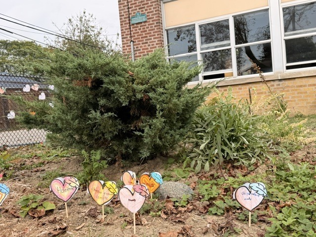 Colorful heart-shaped signs decorate a leafy garden outside a brick school, creating a cheerful, caring feel beneath the windows.