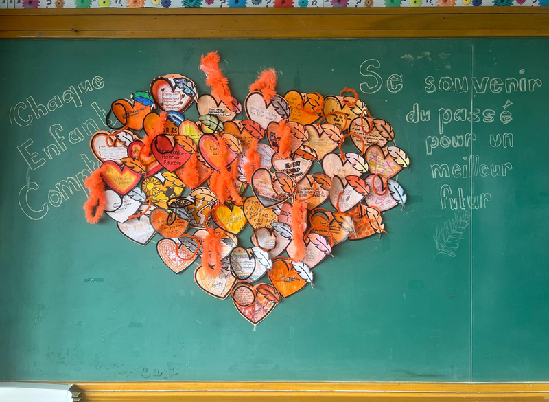 A colorful heart collage of paper hearts and orange feathers decorates a green classroom chalkboard, with French writing about a better future.