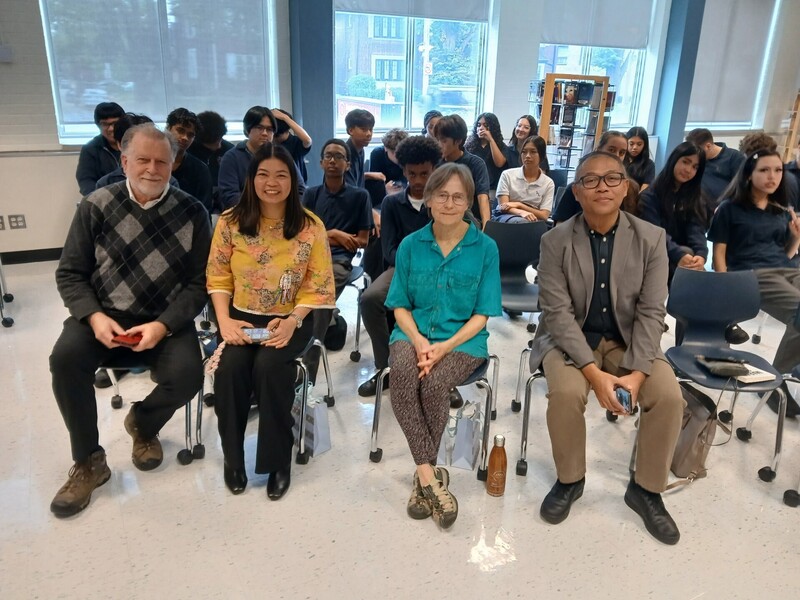 4 adults posing on chairs with students in uniform sitting behind them.