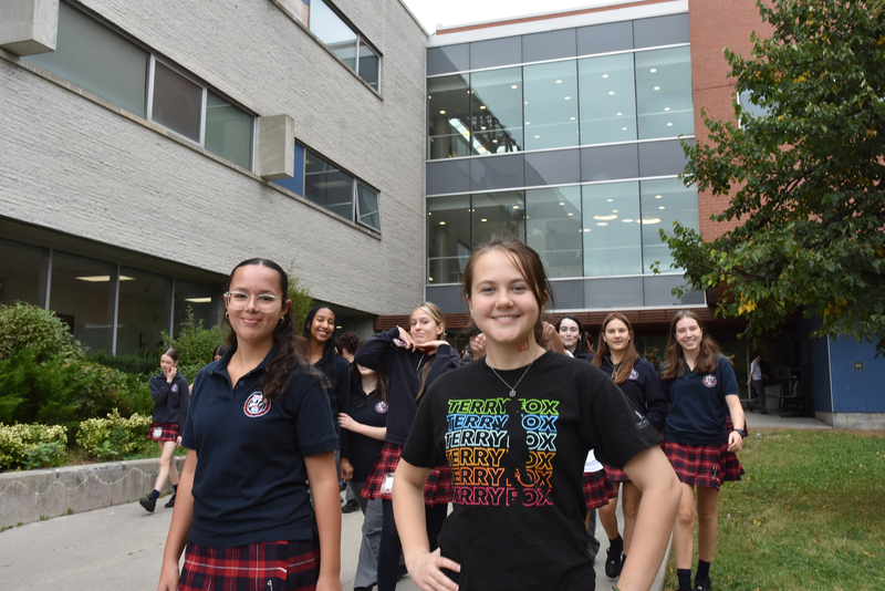 Students in navy polo shirts and plaid skirts walk toward the camera on a campus lawn, with a modern multi-story building and glass windows in the background.