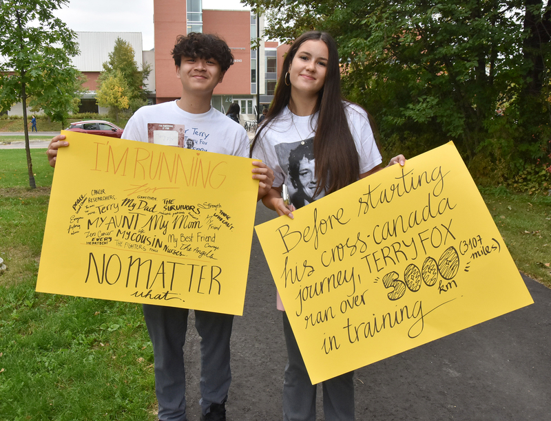 Two people standing outdoors holding bright yellow signs with handwritten messages about their running journey, including “I’m Running,” “No Matter What,” and “Before starting his cross-canada journey, Terry Fox ran over 5,300 miles in training.”