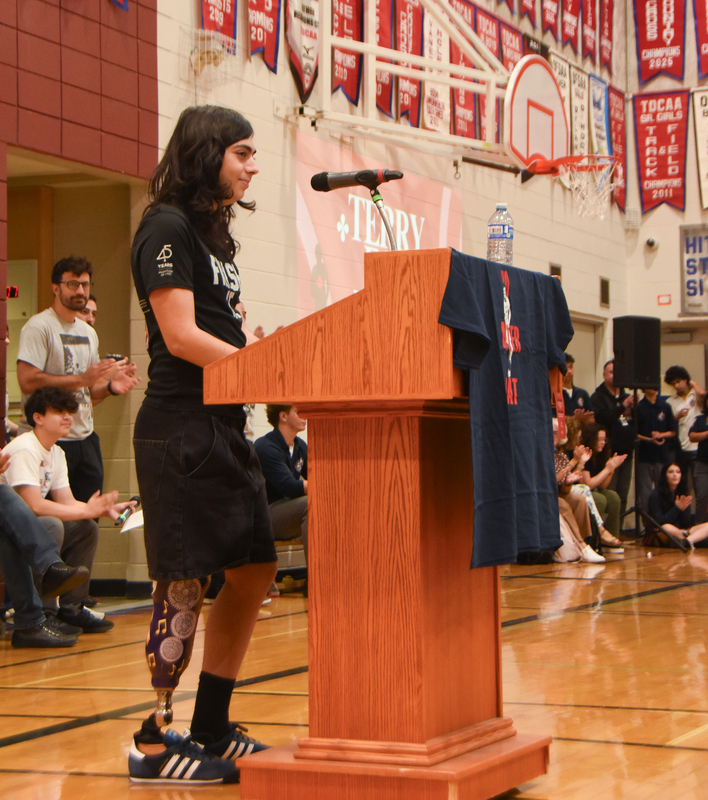 A person in a black outfit speaks at a wooden gymnasium podium with a microphone, while a crowd sits and watches; a basketball hoop and red school banners line the back wall.