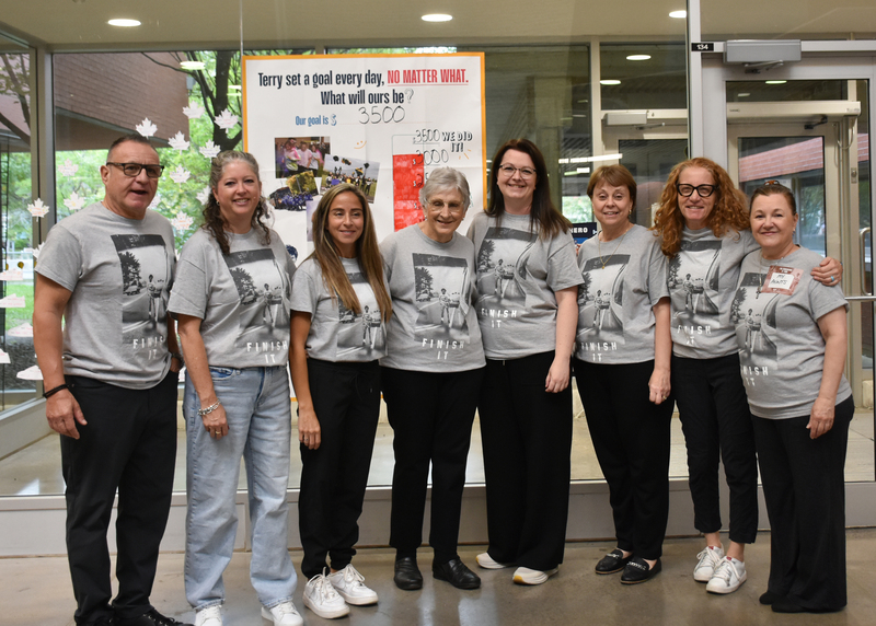 Seven people stand together in matching gray T-shirts in an office lobby, smiling in front of a banner that reads “Terry set a goal every day, NO MATTER WHAT.”