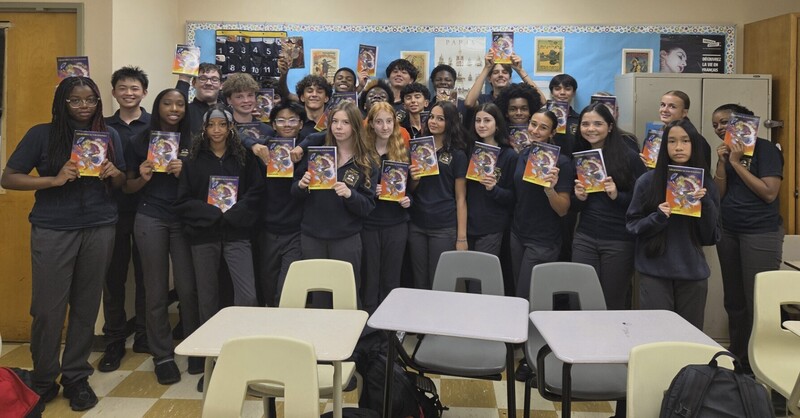 Group of students standing in a classroom holding up printed comic books, with desks and chairs in the foreground and posters on the walls behind them.