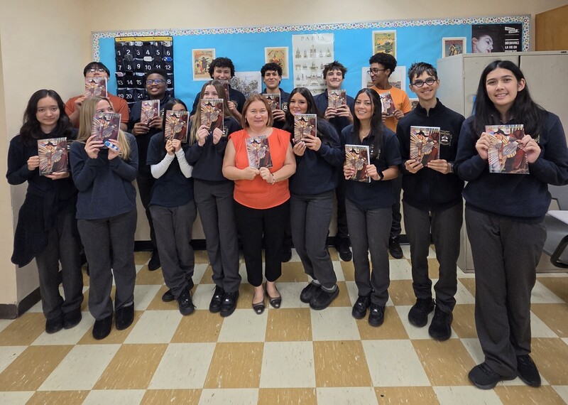 A group of students stand in a classroom holding up printed books or posters, posing for a group photo in front of a bulletin board with colorful displays.