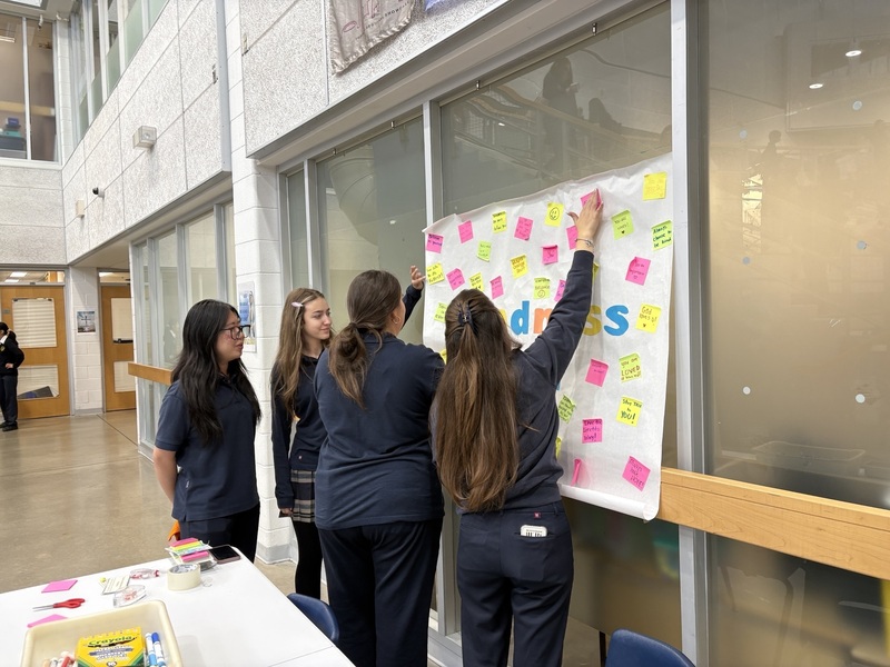 A group of students in blue uniforms gather around a glass wall covered with colorful sticky notes, gesturing as one points to the board during an indoor activity.