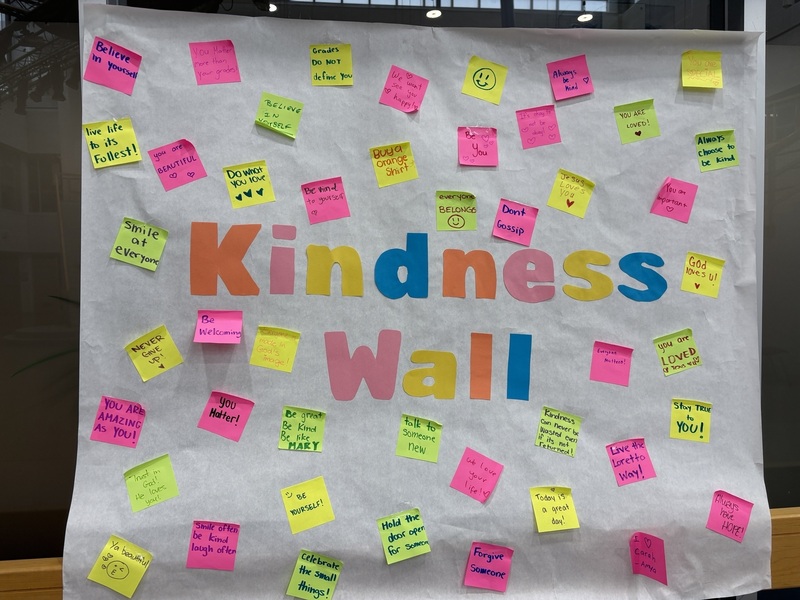 Colorful sticky notes surround the large words “Kindness Wall,” with many small handwritten messages scattered across a light background.