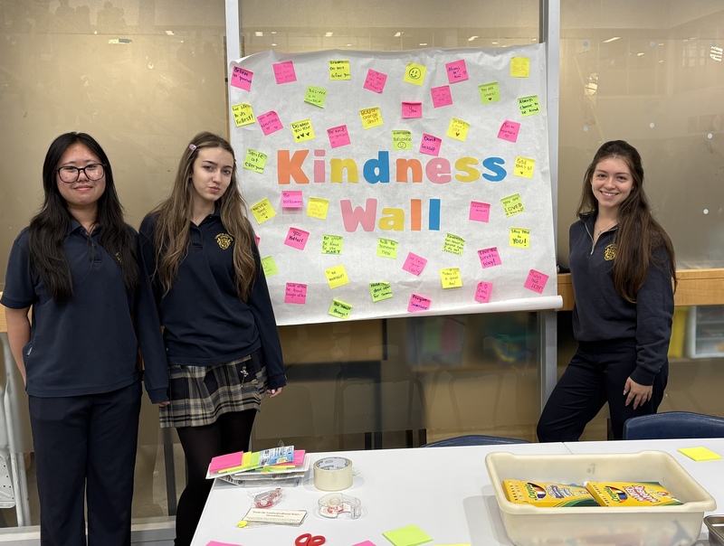 Three people stand in front of a large bulletin board reading “Kindness Wall,” covered with colorful sticky notes, with stationery and a tray of markers on a table in the foreground.