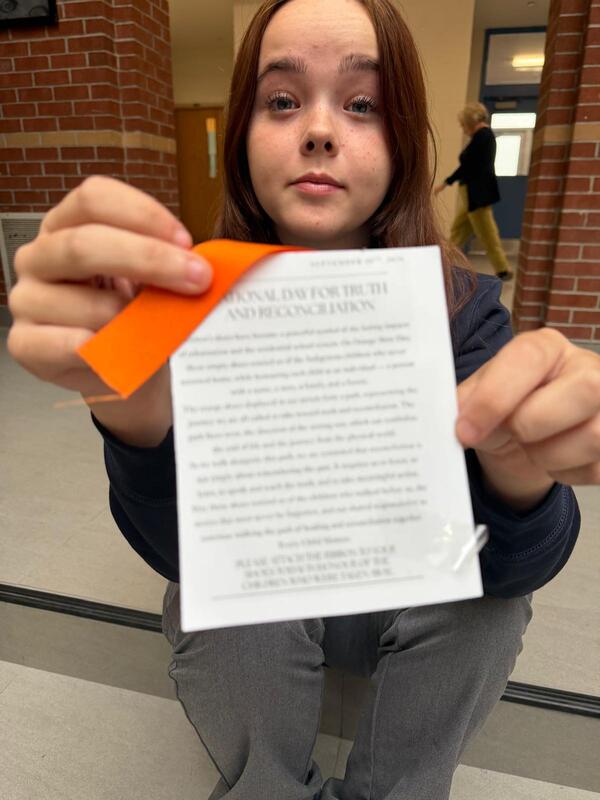 A person holding a printed document close to the camera while a bright orange sticky note is placed on top; brick columns and a corridor with a blurred passerby are in the background.
