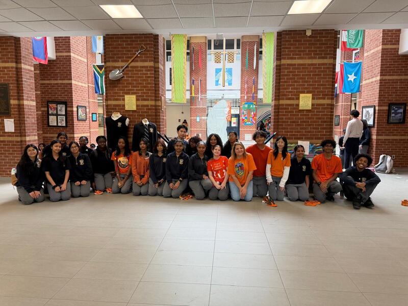 Large group photo in a school hallway with brick walls, flags and framed displays behind, several people standing and kneeling in rows wearing orange shirts and dark tops, with two standing near the center.