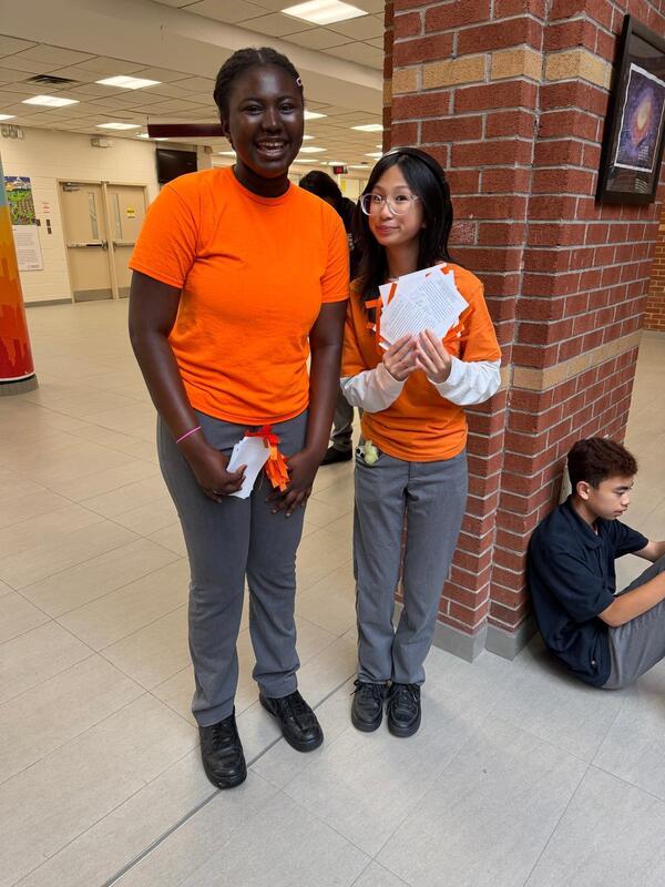 Two people in orange shirts stand in a school hallway beside a brick column; one holds folded papers and the other stands smiling, while a child in dark clothing sits on the floor to the right.