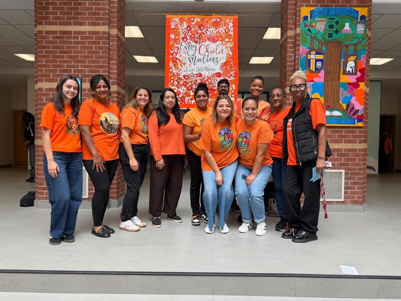 Group of people in orange shirts posing for a photo outside a brick building hallway, standing in front of a red-orange banner and a colorful mural on the wall.
