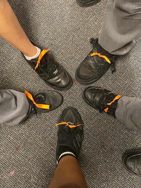 Five pairs of black shoes with bright orange laces are arranged in a circle on a gray carpet, with one person’s legs and hands visible in the center.