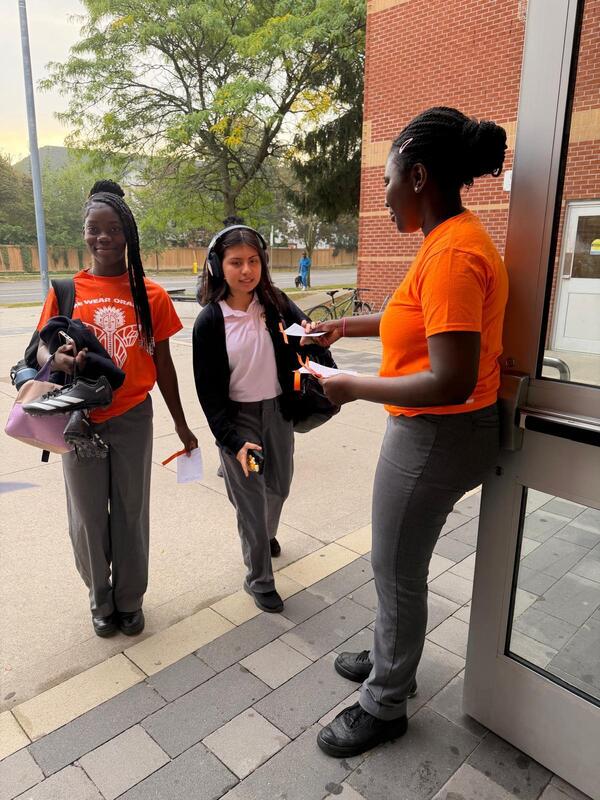 Three students in orange and black outfits stand outside a building, one handing something to another near a doorway while the third watches; trees and a campus walkway are in the background.