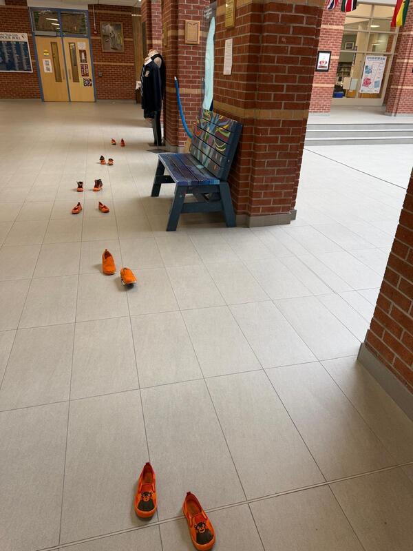 A hallway with brick pillars and tiled floor where several bright orange shoes are scattered in a line and clusters, with a bench and a person standing near the far end.