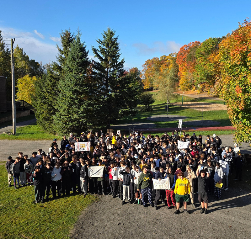 Group photo of Chaminade students and staff at Camp Manitouon a sunny autumn day, holding signs before colorful fall trees