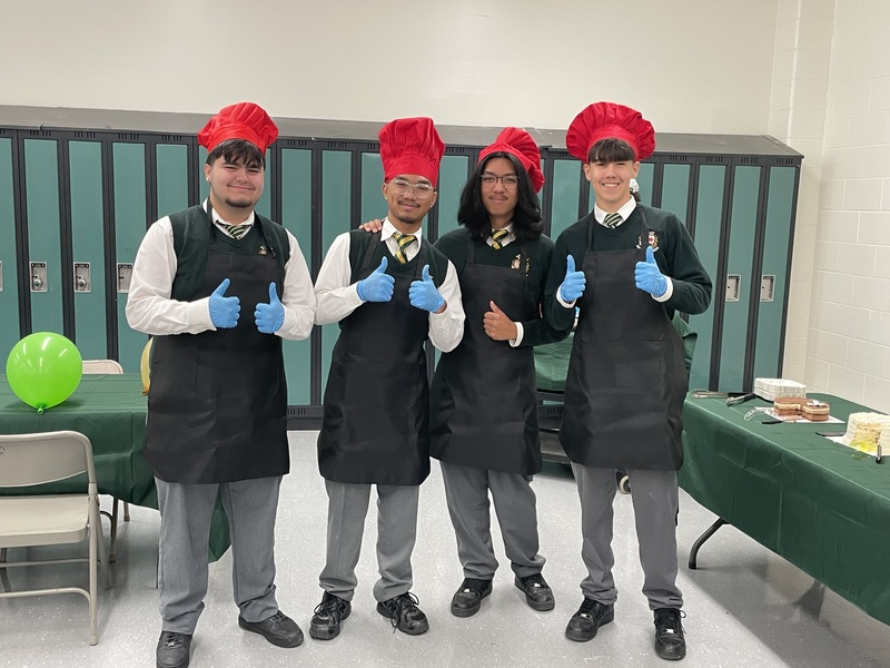 Chefs wearing red tall hats and black aprons stand in a locker-room setting, smiling and giving thumbs-up with blue gloves; green lockers, balloons, and a table with food are in the background.