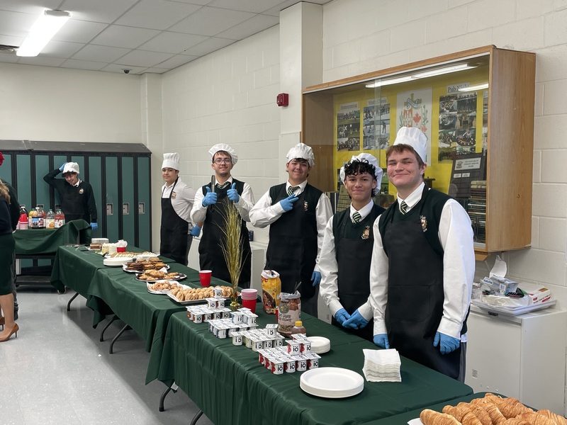 Group of chefs in a break room wearing white and red hats, posing behind a long table of food, giving thumbs-up and smiling in front of windows.