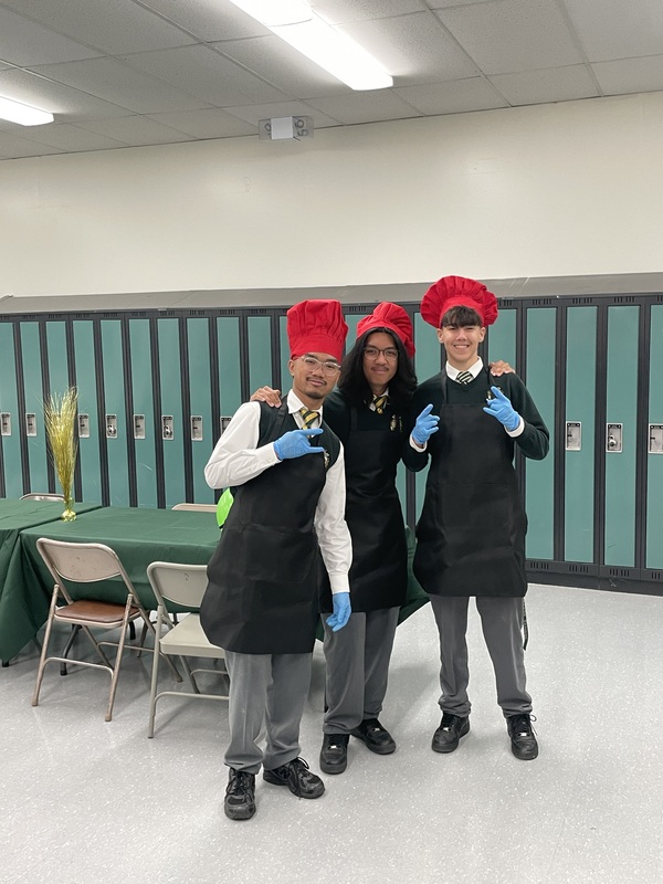 Chefs wearing red tall hats and black aprons stand in a locker-room setting, smiling and giving thumbs-up with blue gloves; green lockers, balloons, and a table with food are in the background.