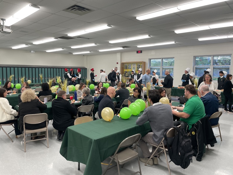 A community event in a bright room with rows of round tables covered in green cloth, attendees seated and standing, and green and yellow balloons decorating the space.