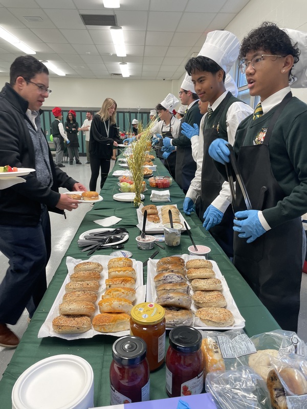 Students in chef hats and aprons stand behind a long table covered with sandwiches, cookies, jars of jam, and other food items in a cafeteria during an event.