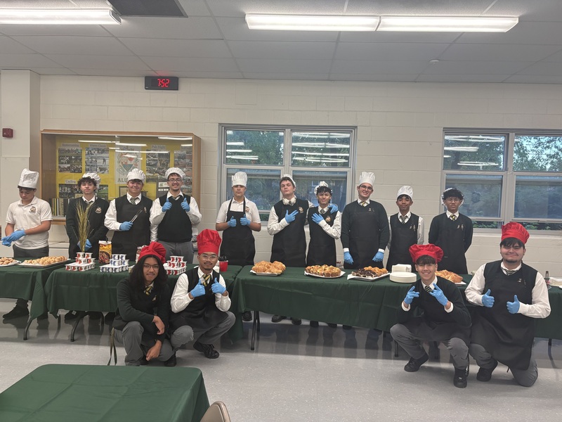 Group of chefs in a break room wearing white and red hats, posing behind a long table of food, giving thumbs-up and smiling in front of windows.