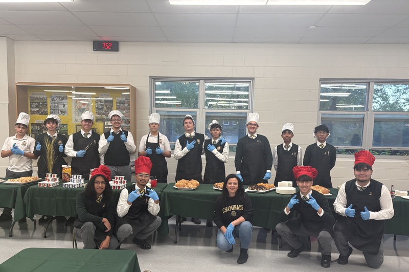 Group of chefs in a break room wearing white and red hats, posing behind a long table of food, giving thumbs-up and smiling in front of windows.