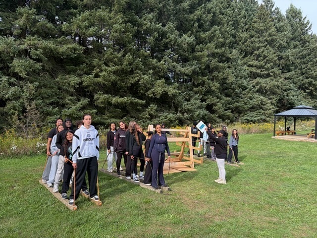 Group of people standing on a grassy lawn near a wooden structure outdoors, with a large evergreen tree line in the background and a small covered pavilion to the right.