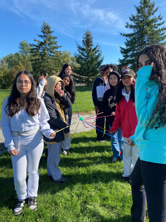 A group of people stand outdoors on a sunny grassy lawn, holding colorful ropes and posing for a photo among tall evergreen trees under a blue sky.