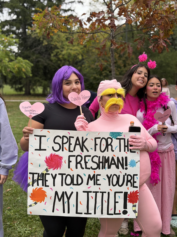 Students in pink and purple clothing pose with a sign that says I speak for the freshman! They told me you're my little!