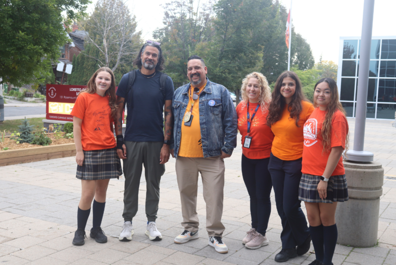 A group of six people wearing orange shirts and casual clothes stand together outdoors on a paved walkway near trees and a building entrance.