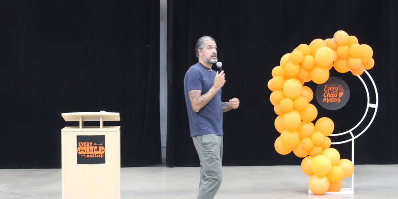 A man speaks on a stage next to a podium and a large balloon number “3” made of orange and yellow balloons, with a black backdrop and gray floor.